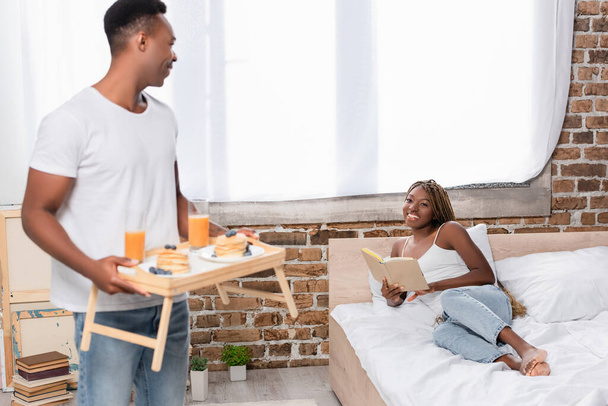 Smiling african american woman with book looking at boyfriend with breakfast on tray on blurred foreground in bedroom  - 写真・画像