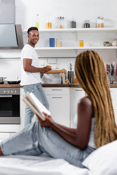 Souriant homme afro-américain tenant des crêpes près du plateau et de la petite amie avec un livre au premier plan flou sur le lit  - Photo, image