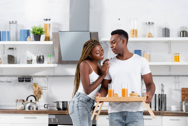 Smiling african american woman hugging boyfriend with delicious breakfast and orange juice on tray in kitchen  - Photo, Image
