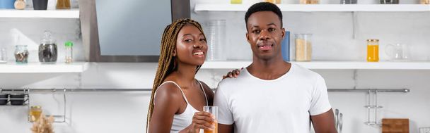Cheerful african american woman holding glass of orange juice and hugging boyfriend in kitchen, banner  - Фото, изображение