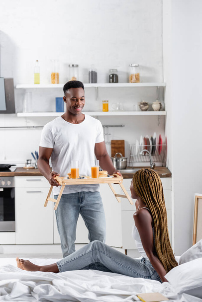 Smiling african american man holding orange juice and pancakes on tray near girlfriend on bed on blurred foreground  - Fotografie, Obrázek