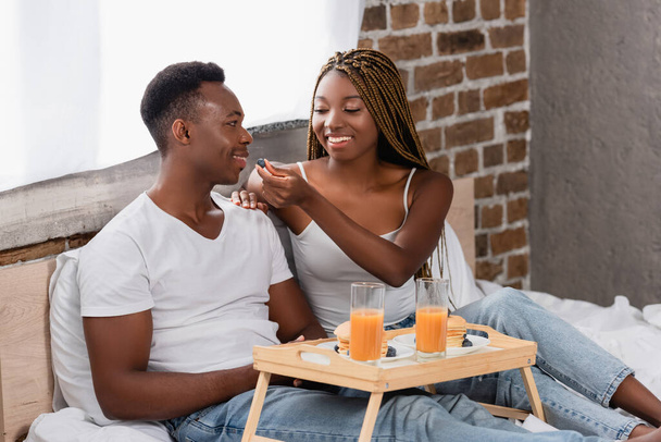 Smiling woman feeding african american boyfriend with blubbery near breakfast on tray on bed   - Foto, afbeelding