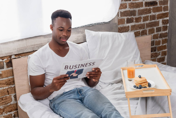 Sonriente hombre afroamericano leyendo periódico de negocios cerca del desayuno en bandeja en la cama  - Foto, imagen