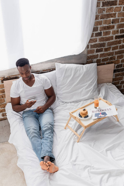 High angle view of african american man using smartphone near breakfast and business newspaper on tray on bed  - Foto, Imagen
