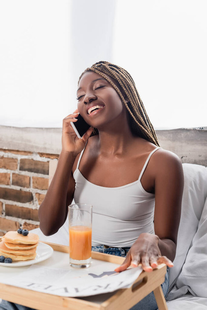Cheerful african american woman talking on smartphone near breakfast and newspaper on tray on bed  - Фото, зображення