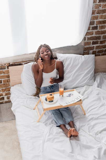 Cheerful african american woman talking on smartphone near breakfast and travel newspaper on bed  - Fotografie, Obrázek