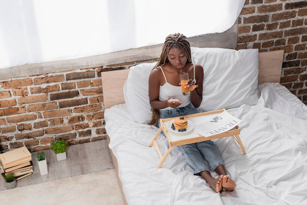 Mujer afroamericana usando smartphone y sosteniendo un vaso de jugo de naranja cerca de periódico de viaje y panqueques en la cama  - Foto, imagen