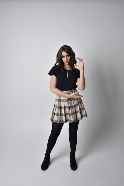 Full length portrait of pretty brunette woman wearing tartan skirt and blouse.  Standing pose on the ground,  against a  studio background. - Foto, afbeelding