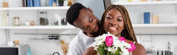 Sonriente mujer afroamericana sosteniendo ramo cerca de novio en la cocina, pancarta  - Foto, imagen