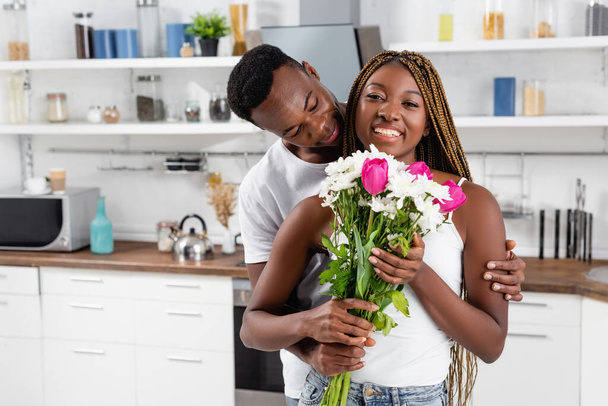 African american man hugging cheerful girlfriend with bouquet in kitchen  - Фото, изображение