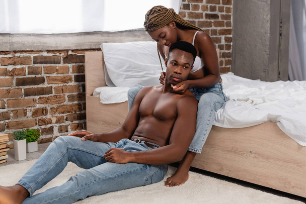 Afrikkalainen amerikkalainen nainen farkut ja rintaliivit koskettaa paidaton mies lattialla makuuhuoneessa  - Valokuva, kuva