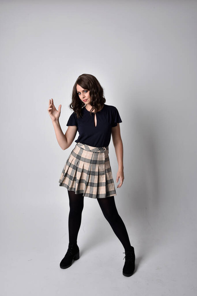 Full length portrait of pretty brunette woman wearing tartan skirt and blouse.  Standing pose on the ground,  against a  studio background. - Foto, imagen
