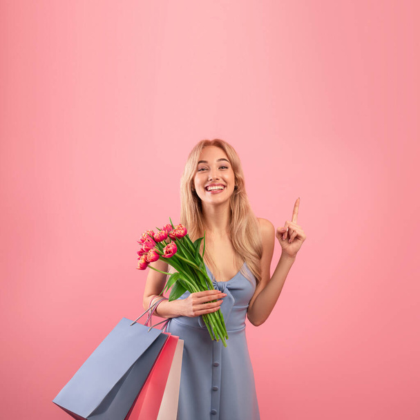 Happy young lady wearing lovely dress, holding spring flowers and gift bags, pointing at empty space on pink background - Photo, Image