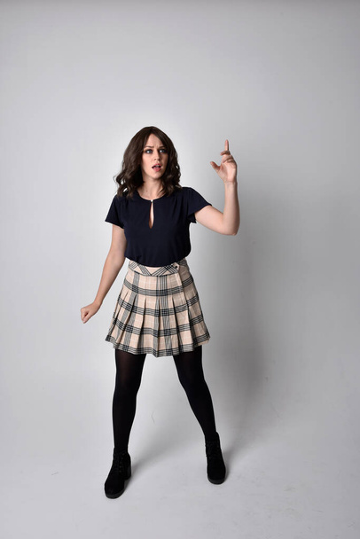 Full length portrait of pretty brunette woman wearing tartan skirt and blouse.  Standing pose on the ground,  against a  studio background. - Photo, Image