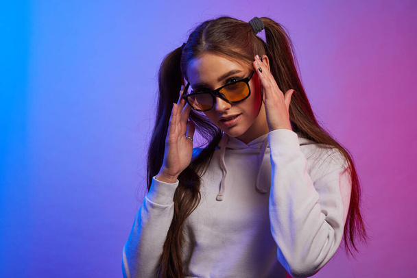 Portrait of young woman wearing trendy yellow lens sunglasses seriously looking straight on camera in neon light. Hipster teenager in glasses. - Fotoğraf, Görsel