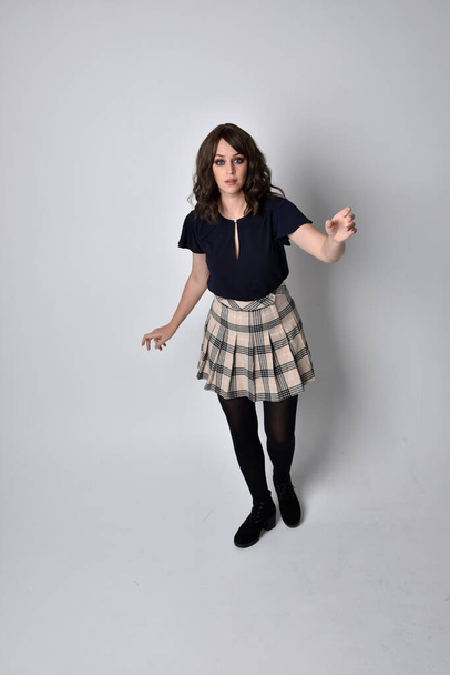 Full length portrait of pretty brunette woman wearing tartan skirt and blouse.  Standing pose on the ground,  against a  studio background. - Foto, imagen