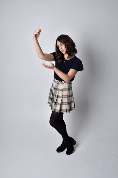 Full length portrait of pretty brunette woman wearing tartan skirt and blouse.  Standing pose on the ground,  against a  studio background. - Photo, Image