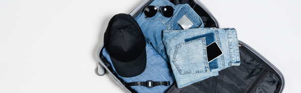 πάνω όψη του κινητού τηλεφώνου στην τσέπη του τζιν κοντά σε πουκάμισο denim, μαύρο καπάκι, ηλεκτρονικό ρολόι χειρός και γυαλιά ηλίου σε βαλίτσα σε λευκό, banner - Φωτογραφία, εικόνα