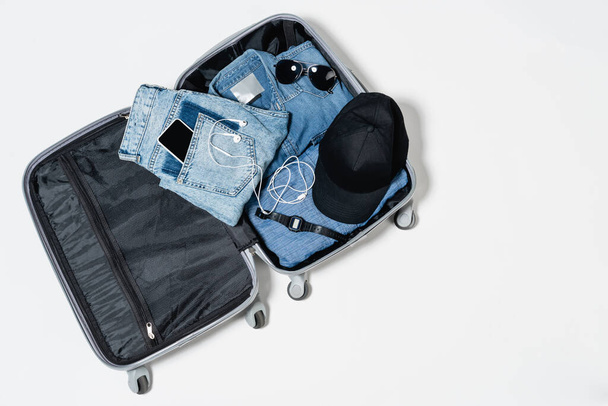 top view of suitcase with denim clothes, mobile phone, black cap, electronic wristwatch, sunglasses and earphones on white - 写真・画像