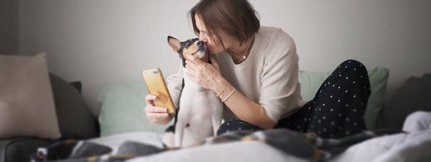 Beautiful young adult woman sitting in pajamas on the bed in the morning kissing pet with bassengi dog while making selfie on smartphone - Fotografie, Obrázek