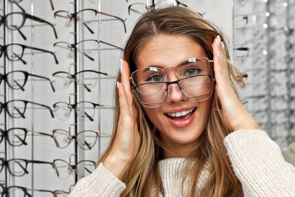 Woman chooses glasses in the store. Blonde in a beige sweater buys glasses. Girl on a background of shop windows with different models of glasses. - 写真・画像