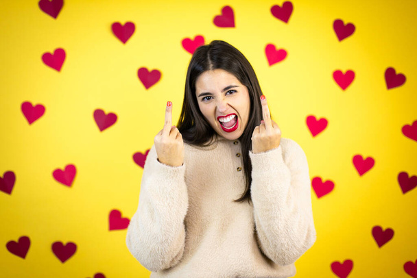 Young caucasian woman over yellow background with red hearts showing middle finger doing fuck you bad expression, provocation and rude attitude. screaming excited - Foto, afbeelding