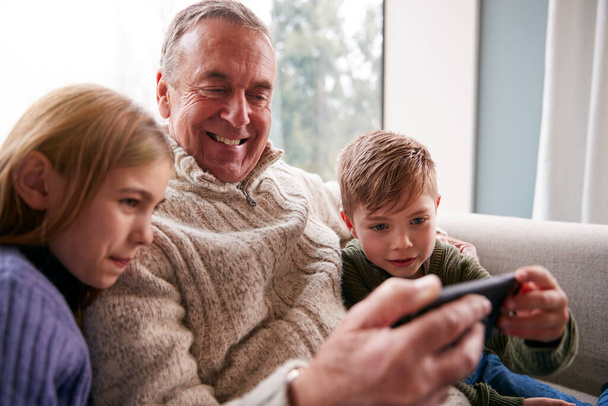 Grandchildren On Sofa At Home Showing Grandfather How To Use Mobile Phone - 写真・画像