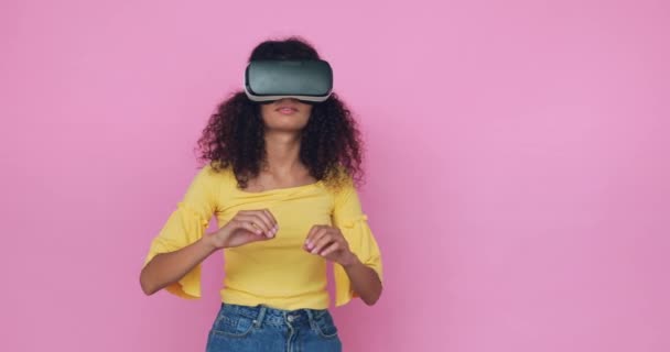 curly young woman in vr headset swiping and gesturing isolated on pink - Footage, Video