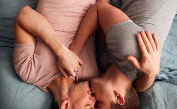 Overhead Shot Of Loving Same Sex Male Couple Lying On Bed At Home Hugging And Kissing Together - Foto, immagini