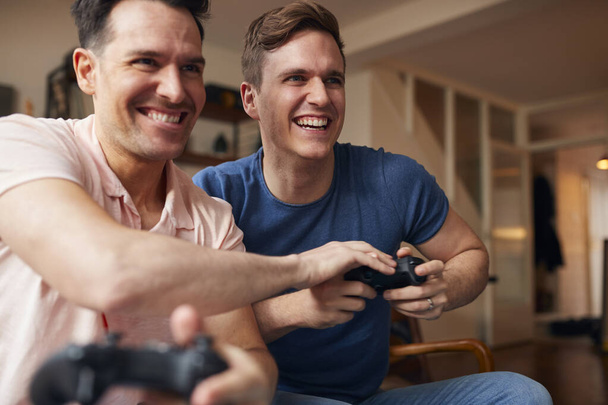 Loving Same Sex Male Couple Sitting On Sofa At Home Gaming Together - Фото, изображение