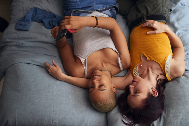 Overhead Shot Of Loving Same Sex Female Couple Lying On Bed At Home Looking At Mobile Phone Together - Fotografie, Obrázek