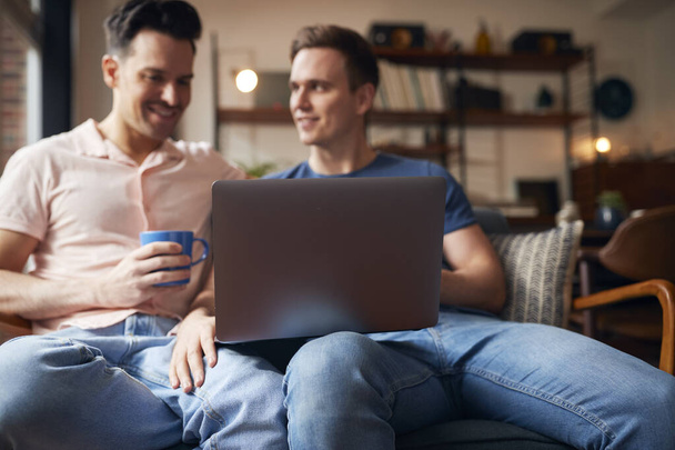 Loving mismo sexo masculino pareja sentado en sofá usando portátil como ellos relajarse en casa juntos - Foto, imagen