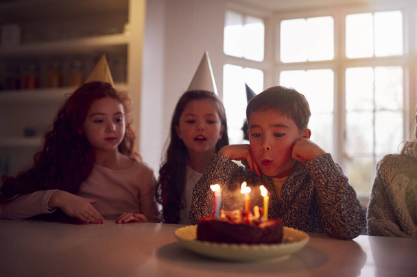 Chłopiec zdmuchując świece na ciasto, jak świętuje urodziny z grupą przyjaciół w domu - Zdjęcie, obraz