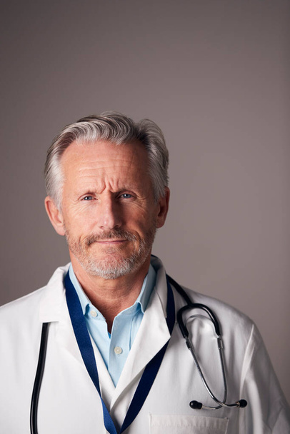Studio Portrait Of Mature Male Doctor Wearing White Coat Standing Against Grey Background - Foto, imagen