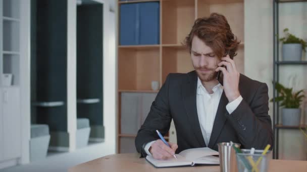 Μέτριο πλάνο του όμορφου Καυκάσου επιχειρηματία που φοράει κοστούμι κάθεται στο χώρο εργασίας, έχει τηλεφωνική συνομιλία με τον επιχειρηματικό συνεργάτη και κρατώντας σημειώσεις στο σημειωματάριο - Πλάνα, βίντεο