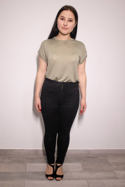 okouzlující brunetka žena v bažinaté barvě tričko a černé kalhoty na šedém pozadí stěny. herečka, emoce. kopírovat prostor  - Fotografie, Obrázek