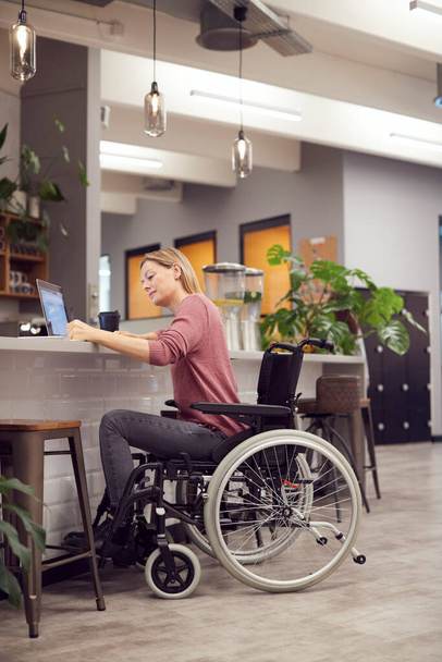 Businesswoman In Wheelchair Working On Laptop In Kitchen Area Of Busy Modern Office - Photo, image