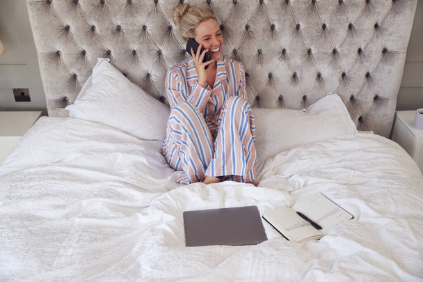 Businesswoman In Pyjamas Sitting On Bed Making Call On Mobile Phone Working From Home - Photo, Image