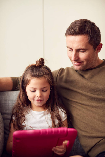 Pai e filha sentados no sofá em casa brincando juntos no tablet digital no caso rosa em casa - Foto, Imagem