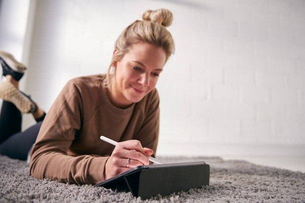 Woman Drawing On Digital Tablet Using Stylus Pen Lying On Carpet At Home - 写真・画像