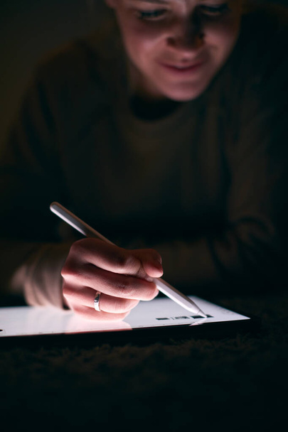 夜間カーペットの上に横たわるスタイラスペンを使用したデジタルタブレットで描く女性のクローズアップ - 写真・画像