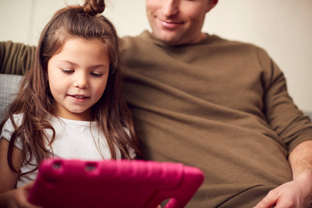 Father And Daughter Sitting On Sofa At Home Playing Together On Digital Tablet In Pink Case At Home - Фото, изображение