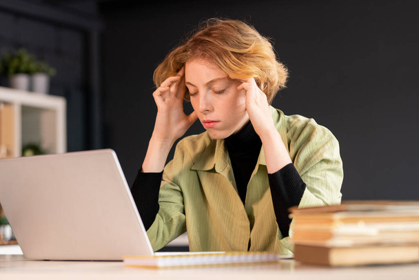 Junge blonde Softwareentwicklerin sitzt am Schreibtisch und versucht sich zu konzentrieren, während sie während des Netzwerks den Kopf vor dem Laptop berührt - Foto, Bild