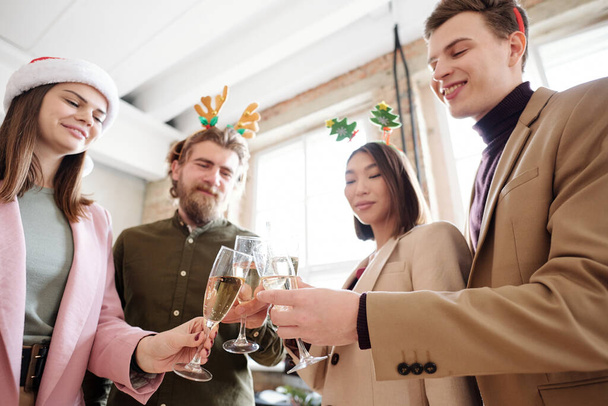 Gruppe junger, gut gelaunter Manager in schicker Freizeitkleidung und weihnachtlichem Stirnband, die am Weihnachtstag mit Champagnerflöten klirren - Foto, Bild