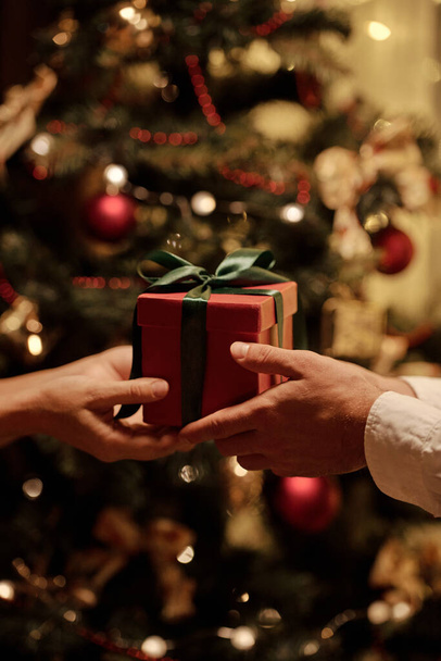 Manos de mujer joven dando caja de regalo llena de rojo atada con cinta verde oscuro a su marido contra el árbol de Navidad decorado - Foto, imagen