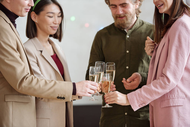 Happy colleagues in smart casualwear clinking with flutes of champagne while toasting for new business projects and Christmas at party - Photo, Image