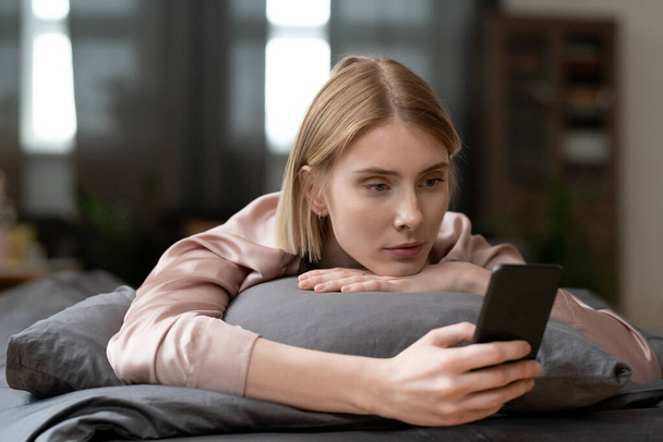 Young blond female in beige silk robe or pajamas making selfie or scrolling through online news in the morning while relaxing on bed after sleep - Fotografie, Obrázek