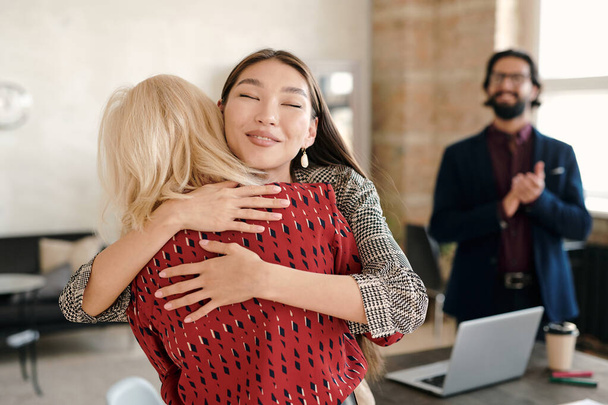 Feliz mujer de negocios asiática felicitando a uno de sus colegas por su gran trabajo mientras la abraza contra un hombre joven y elegante aplaudiendo - Foto, imagen