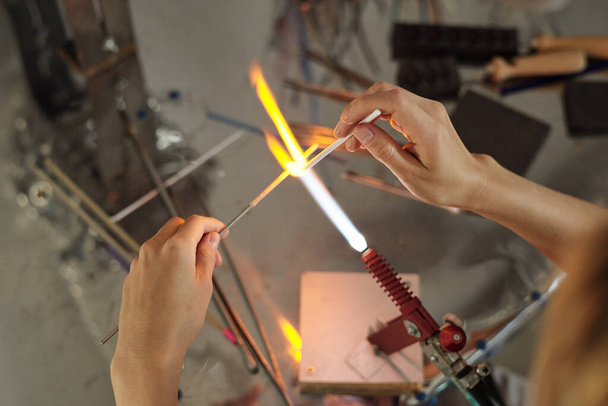 Handen van jonge ambachtelijke vrouw zitten door werkplek in studio en het verbranden van twee stokvormige werkstukken in brander, terwijl het in brand - Foto, afbeelding