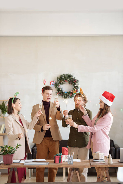 クリスマスプレゼントとテーブルに立っている間、彼らの手の中にベンガルライトを燃焼見てシャンパンのフルートを持つ興奮した若い同僚 - 写真・画像
